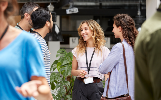Curly-haired woman with lanyard smiling as she chats with two colleagues in a modern workspace