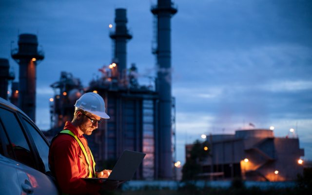 A worker in a hard hat uses a laptop near an industrial plant at twilight.