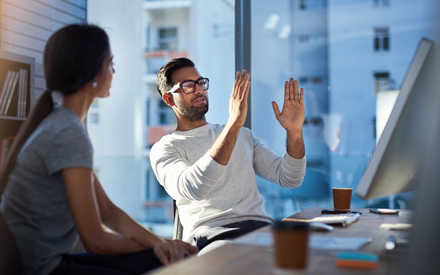 Bearded man with glasses animatedly explains work to female colleague at office desk