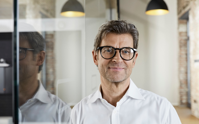 A middle-aged man with glasses smiles, standing in a modern office space.