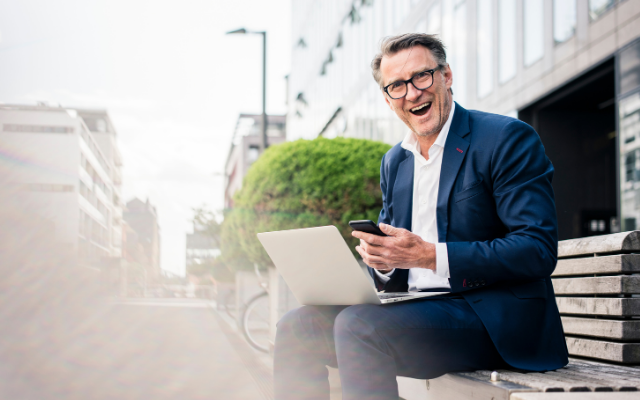 Man in a suit smiling while using a laptop and phone on a bench in an urban setting.