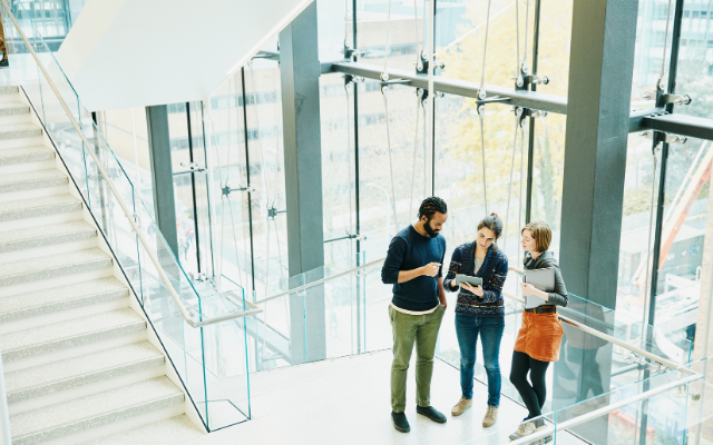 Three people discuss plans on a staircase in a modern, glass-walled building.