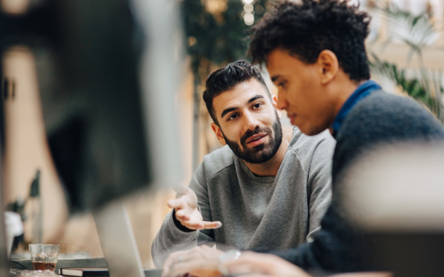 Two men engaged in a discussion at a desk, one gesturing while the other listens intently.