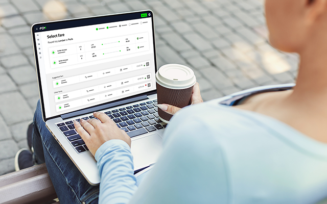 Person outdoors typing on laptop showing a green stock chart, holding a coffee cup