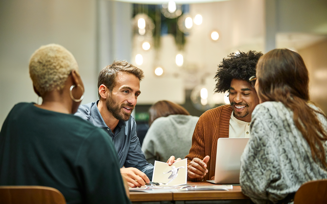 Photo of four diverse coworkers chatting around a laptop at a cafe-style office table