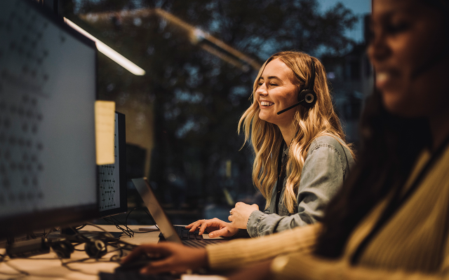 Photograph of a smiling woman wearing headphones working at a computer in a dimly lit office.