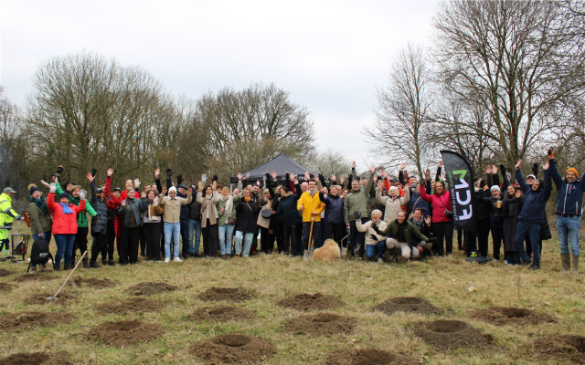 Outdoor group of about 60 people and a dog waving on a grassy field with bare trees.