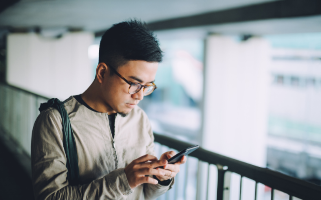 Photograph of a young man with glasses and backpack checking his smartphone on indoor walkway