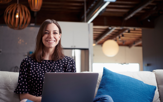 Smiling woman with long brown hair sitting on a couch, working on a laptop.