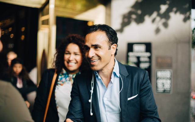 Smiling man in a suit converses with a woman outside a building, with warm lighting.