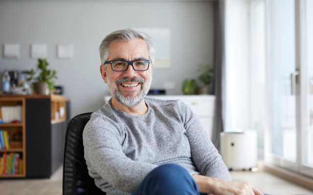 Smiling older man with glasses sitting in a modern room, sunlight streaming in.
