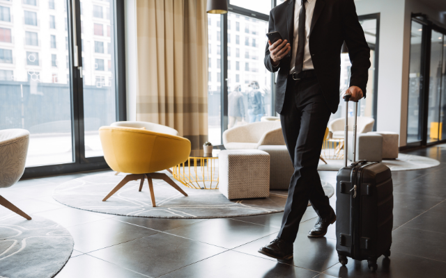 A man in a suit walks through a modern lobby with a suitcase, checking his phone.