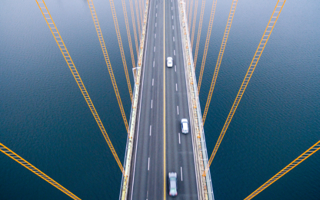Aerial view of a bridge with yellow cables, vehicles moving on a wide road over calm water.