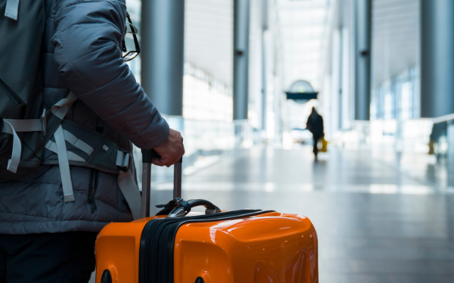 Photograph: traveler in winter coat holding bright orange suitcase in airport terminal corridor