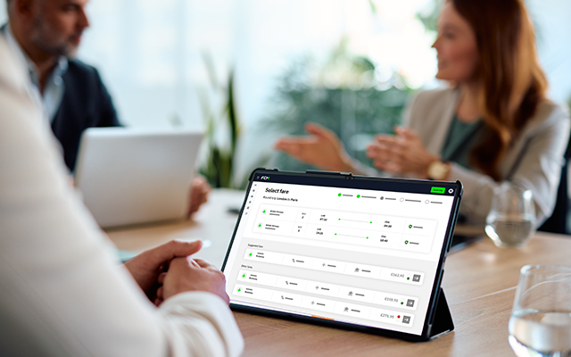 A tablet displaying a dashboard sits on a meeting table, with two professionals discussing.