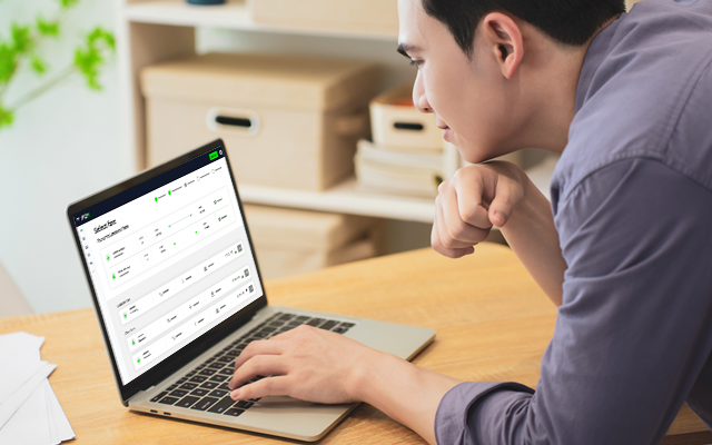 A man sits at a wooden desk, focused on a laptop screen displaying data.