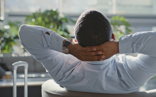 Photograph of man in white shirt leaning back at desk with laptop, hands behind head, gazing out window.