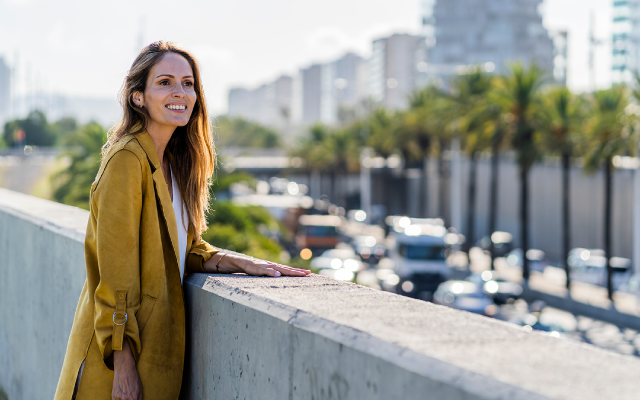 Young woman in mustard coat leaning on a barrier overlooking busy street with palm trees.