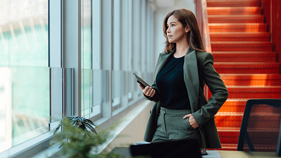 Woman in green blazer holding phone and briefcase, gazing out office window by orange stairs