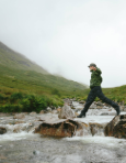 Person in green rain jacket stepping across rocks in a shallow mountain stream under foggy hill