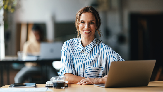 Photograph of a smiling woman in a striped shirt seated at a café desk with laptop and coffee
