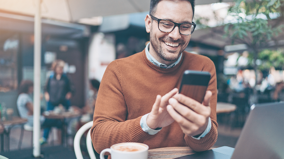 Person in brown sweater scrolling smartphone at café table with coffee cup and laptop