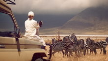 Man in safari truck photographing zebras on a grassy plain with distant mountains