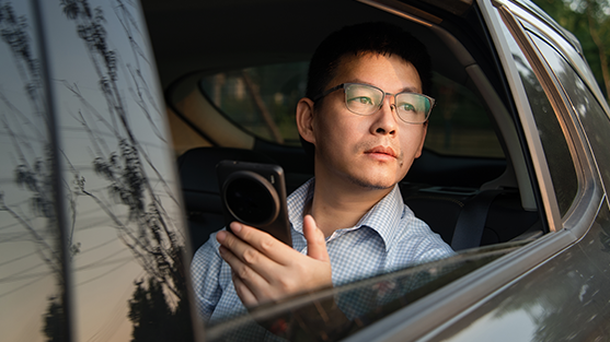 Person with blurred face in car holding camera, trees reflected in window.