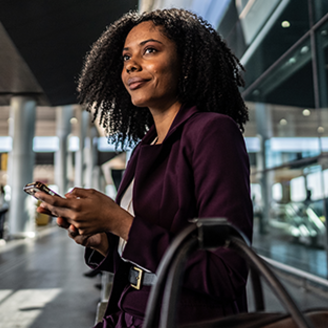 Woman in a purple outfit holding a phone, smiling at an airport terminal.